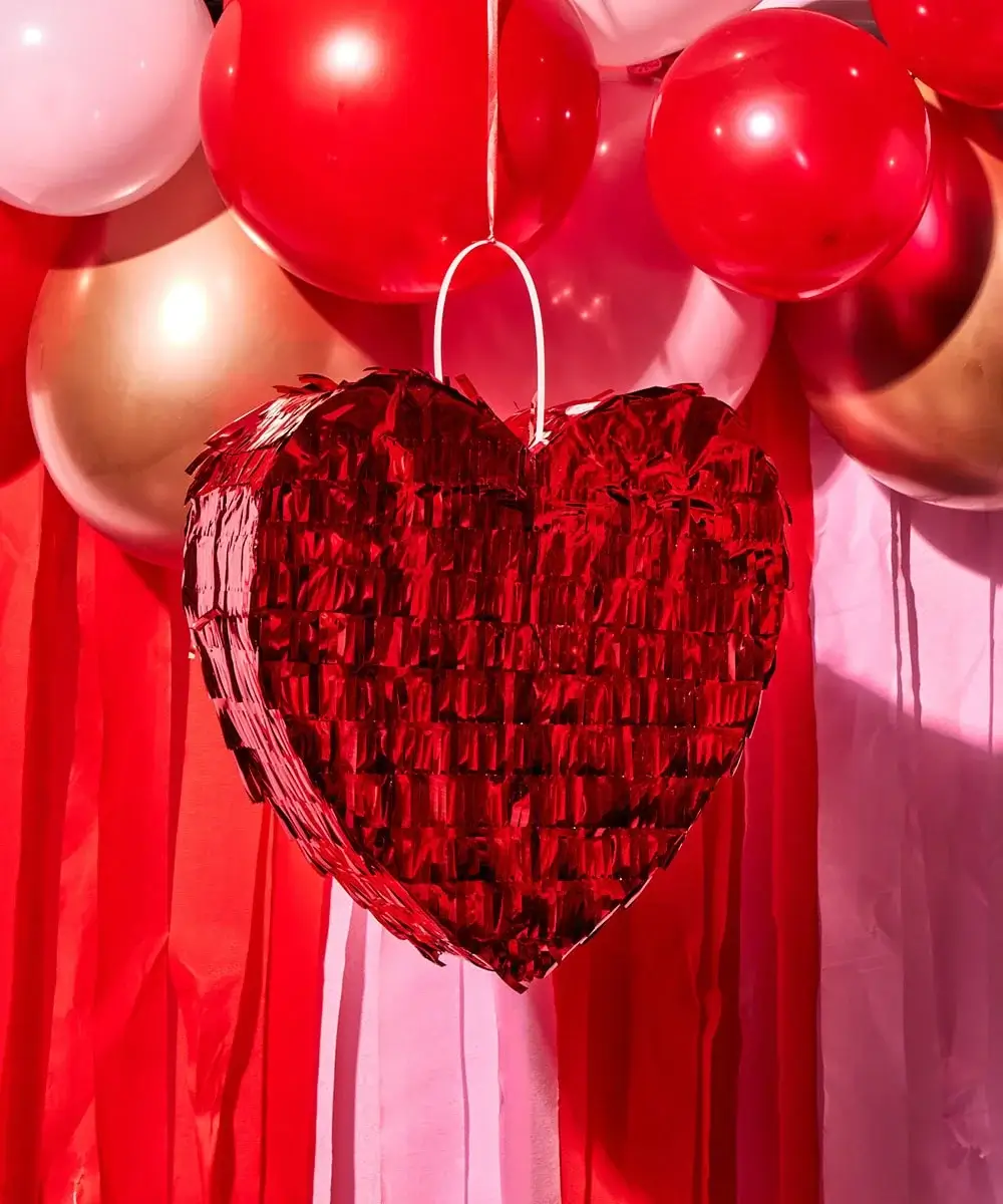 Red heart-shaped piñata hanging in front of red, pink, and gold balloons with matching streamers in the background.